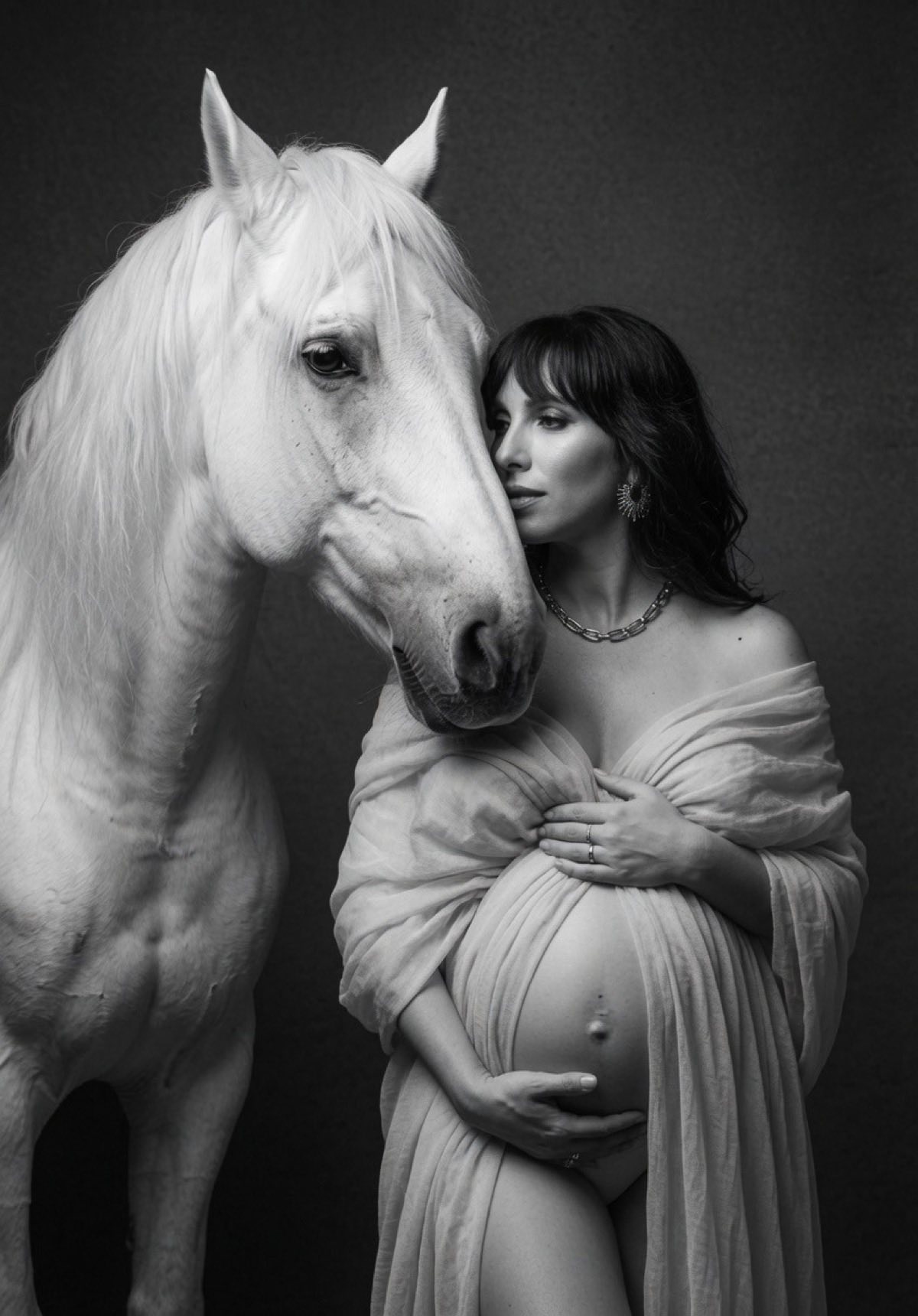 Black-and-white pregnancy portrait of a woman standing beside a white horse in a Chicago studio